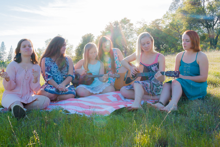 Young Girls Sitting Together in Grassy Field Singing and Playing Musical Instruments With Sunlight Overheadの写真素材