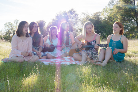 Young Girls Sitting Together in Grassy Field Singing and Playing Musical Instruments With Sunlight Overheadの写真素材