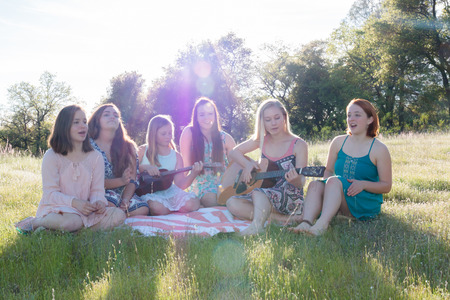 Young Girls Sitting Together in Grassy Field Singing and Playing Musical Instruments With Sunlight Overheadの写真素材