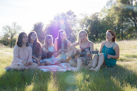 Young Girls Sitting Together in Grassy Field Singing and Playing Musical Instruments With Sunlight Overheadの写真素材
