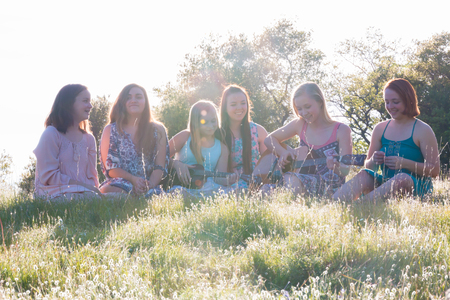 Young Girls Sitting Together in Grassy Field Singing and Playing Musical Instruments With Sunlight Overheadの写真素材