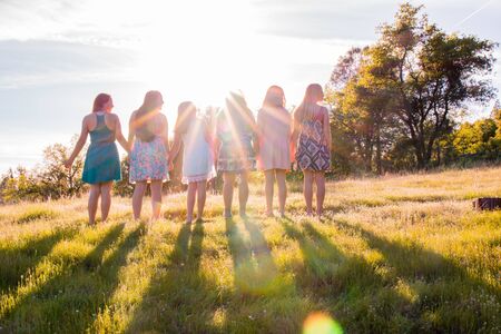 Young Girls Standing Together in Grassy Field Facing the Bright Sunsetの写真素材