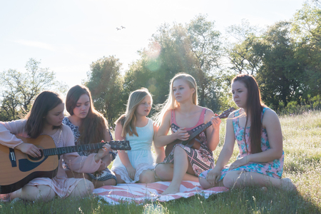 Young Girls Sitting Together in Grassy Field Singing and Playing Musical Instruments With Sunlight Overheadの写真素材