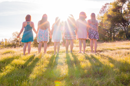 Young Girls Standing Together in Grassy Field Facing the Bright Sunsetの写真素材