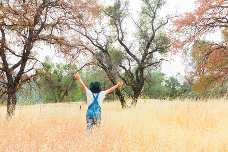 Young Girl With Overalls and Black Hat Standing in Field With Arms Raisedの写真素材