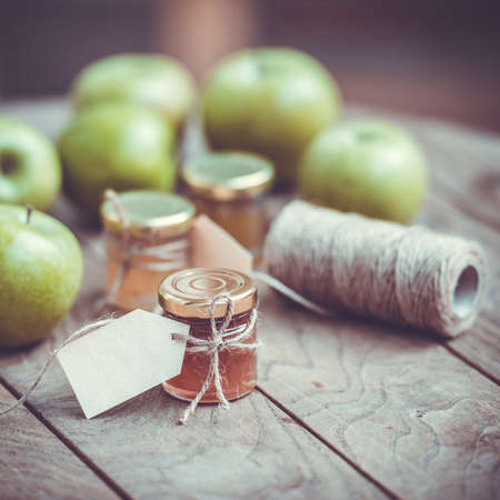 Apple jam in a small glass jar and green apples on wooden table. Blank label provides copy space for a message. Toned imageの写真素材