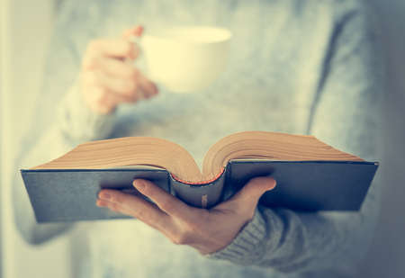 Young woman reading a book and holding cup of tea or coffee. Toned imageの写真素材