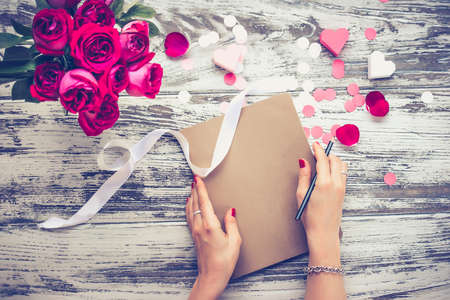 Bouquet of roses and closed book or notebook in female hands on old wooden table. Top view. Toned...