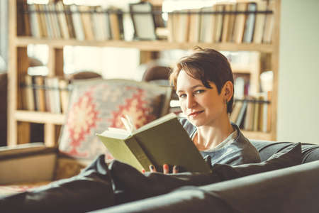 Young beautiful woman reading a book on sofa. Toned picture. Selective focus and shallow DOFの写真素材