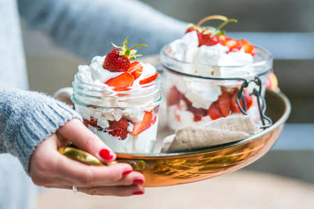 Hands holding classic british summer dessert called Eton Mess. Strawberries, crushed meringue and whipped cream in jarの写真素材