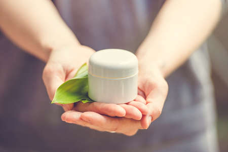 Cosmetic cream container with green herbal leaves in woman hands. Toned imageの写真素材