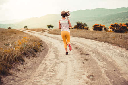 Beautiful girl running on a rural road during sunsetの写真素材