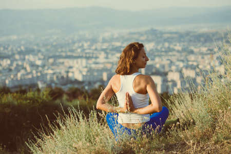 Young woman sits in namaste yoga pose with city on background. Freedom concept. Calmness and relax, woman happiness. Toned imageの写真素材