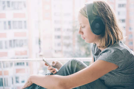 A teenage girl sits near window listening to music and looking at her smartphone. Toned imageの写真素材