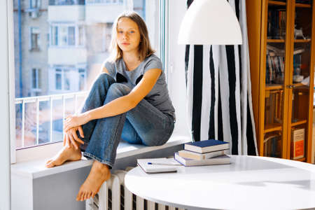 A teenage girl sits on window sill instead of reading books on the tableの写真素材