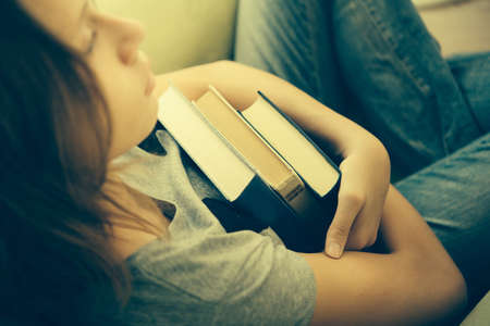 A sad teenage girl sits in armchair holding books. Toned imageの写真素材