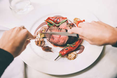Person cuts beef steak with grilled vegetables served on white plate. Toned pictureの写真素材