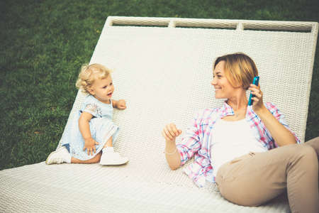 Busy mother is speaking on her smartphone and daughter sitting near her. Toned imageの写真素材