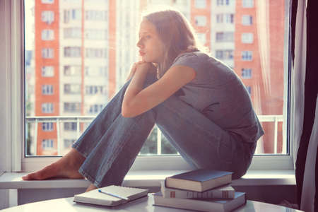 A sad teenage girl sits on window sill instead of reading books on the table. Toned imageの写真素材