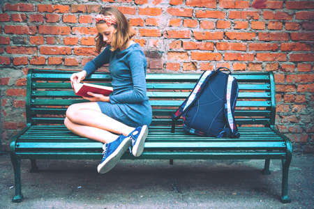 A teenage girl is reading on a bench with brick wall in the background. Toned image