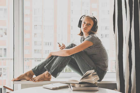 A teenage girl sits on window sill listening to music and looking at her smartphone. Toned imageの写真素材