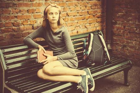 A portrait of a teenage girl sitting on a bench holding a book with brick wall in the background. Toned imageの写真素材