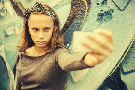 A teenage girl takes selfie with graffiti in the background. Toned image