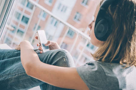 A teenage girl sits near window listening to music and looking at her smartphone. Toned imageの写真素材