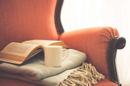 Book, cup and warm plaid on cozy retro armchair. Reading and home decoration concept. Toned pictureの写真素材