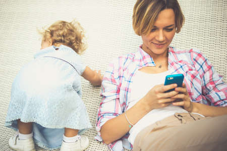 Young beautiful mother checks messages on her smartphone while her daughter has fun. Toned imageの写真素材