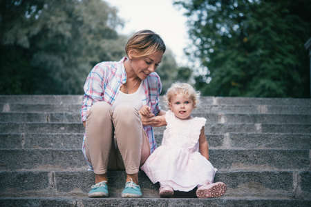 Blonde beautiful mother and daughter sit at the stairs during a walk outdoorsの写真素材