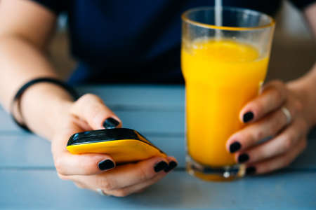 Woman is drinking orange juice with smartphone in her handの写真素材