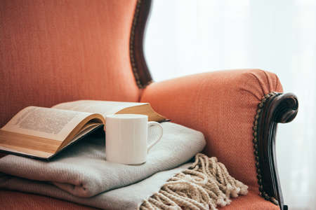 Book, cup and warm plaid on cozy retro armchair. Reading and home decoration conceptの写真素材