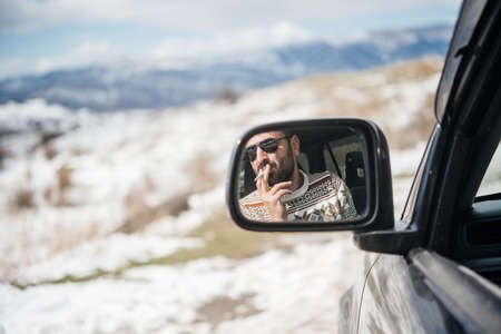 Young man driving the car is seen reflected in the side mirror with mountains in the background. Travel and adventure concept.の写真素材