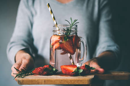 Hands holding retro glass jar of lemonade or cocktail with strawberry and rosemary on wooden board. Toned pictureの写真素材
