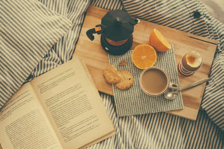 Breakfast served in bed - boiled egg, fresh orange, cookies and coffee, while reading a book. Selective focus. Toned pictureの写真素材