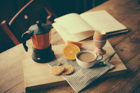 Breakfast served on wooden table - boiled egg, fresh orange, cookies and coffee, while reading a book. Selective focus. Toned pictureの写真素材