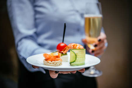 A person holds in hands a plate with different snacks and appetizers and a glass of champagne at a cocktail party. Catering concept.の写真素材