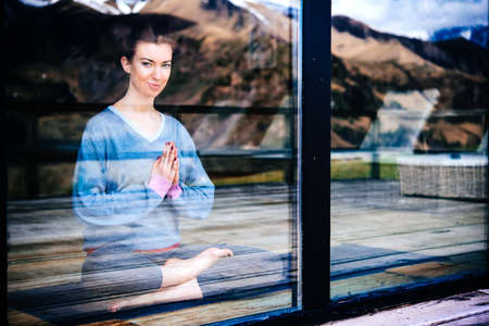 Young beautiful woman meditating while practicing yoga with mountain view in the window reflection. Freedom concept. Calmness and relax, woman happiness.の写真素材