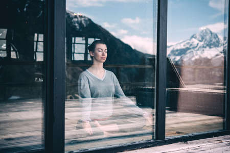 Young beautiful woman meditating while practicing yoga with mountain view in the window reflection. Freedom concept. Calmness and relax, woman happiness. Toned pictureの写真素材