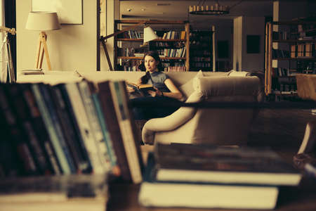 Young beautiful woman enjoys reading sitting on a comfortable couch. Learning and knowledge concept. Toned pictureの写真素材