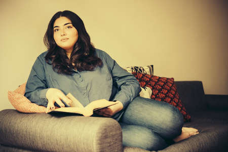 Beautiful plus size girl sitting on sofa reading a book. Toned pictureの写真素材