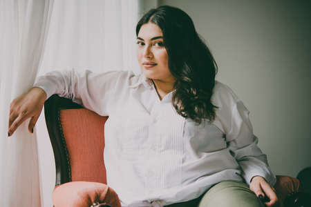 Portrait of beautiful plus size girl in white sitting in the armchair. Toned pictureの写真素材