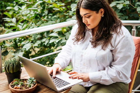Portrait of beautiful plus size girl working on her laptop on balcony or terrace of cafeの写真素材