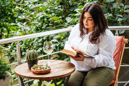 Portrait of beautiful plus size girl reading a book on balcony or terrace of cafeの写真素材