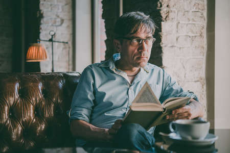 Mature handsome man looking at his smartphone while reading a book in a cafe. Toned picture