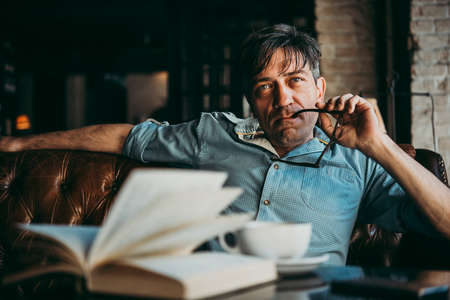 Portrait of a mature confident handsome man while thinking. Book and cup of coffe in frontの写真素材