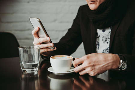 Man holding smartphone and cup of cappuccino coffee in cafeの写真素材