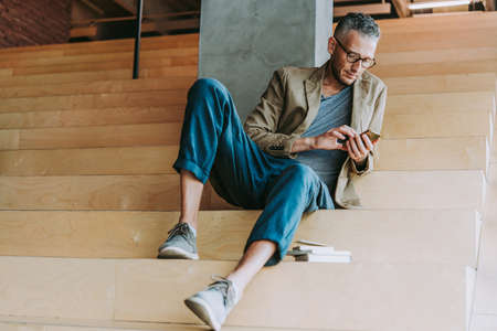 Mature handsome man sitting at the steps and using his smartphone