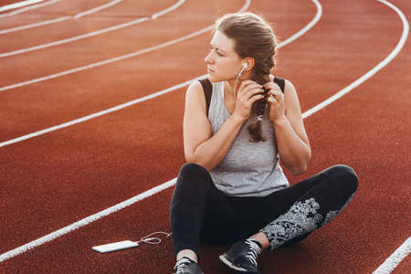 A young beautiful woman having rest with her smartphone sitting at a stadium running track.